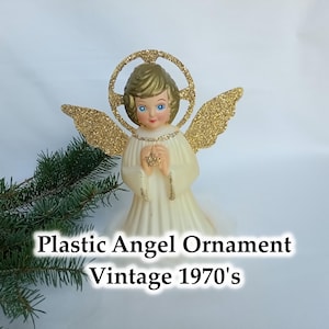 May include: A vintage plastic angel ornament from the 1970s. The angel features a cream dress, gold glitter wings and halo, and blue eyes. The text "Plastic Angel Ornament Vintage 1970's" is displayed below the angel.