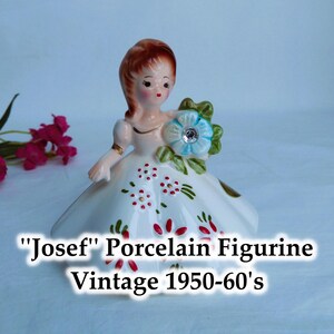 May include: A porcelain figurine of a girl in a white dress with red and green floral accents. She is holding a blue flower with a rhinestone centre. The figurine is labelled "Josef" Porcelain Figurine Vintage 1950-60's.