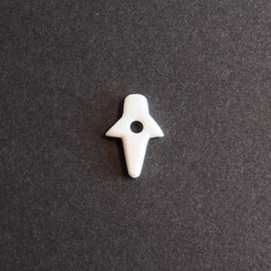 May include: A small, white, cross-shaped object with a circular hole in the center. The object has a smooth, glossy finish and is set against a dark gray background. The design is simple and minimalist.