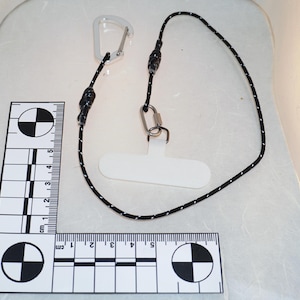 May include: A black and white lanyard with a clear plastic carabiner clip and a silver metal clasp. The lanyard has a white plastic phone holder. A ruler with centimeter markings is in the image.