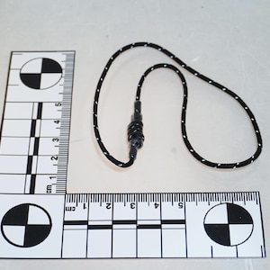 Puede incluir: Un cordón con estampado en blanco y negro con un cierre de plástico negro. El cordón mide aproximadamente 30 cm de largo. Una regla blanca y negra está en el fondo, con medidas en centímetros.