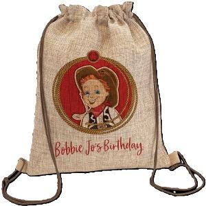 May include: A beige drawstring backpack with brown straps and accents. The front features a circular design with a cartoon image of a cowboy and the text "Bobbie Jo's Birthday" in red.