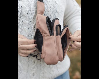 Può includere: Una borsa a tracolla color cipria con più scomparti con cerniera. La borsa ha una tracolla nera e una fodera interna nera. Le cerniere sono nere. La borsa viene tenuta aperta per mostrare l'interno.