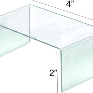 May include: A clear acrylic display stand, shaped like a U, with dimensions of 4 inches wide and 2 inches high. The stand is transparent, allowing for a clear view of items placed upon it. Ideal for showcasing small products.