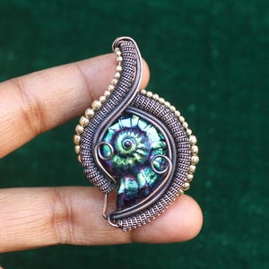 Puede incluir: Un colgante envuelto en alambre con una amonita iridiscente verde y púrpura. El colgante está hecho con alambre de cobre y oro y tiene un diseño delicado e intrincado.