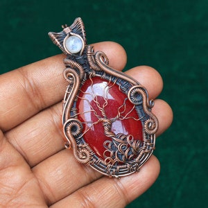 May include: A copper wire wrapped pendant featuring a red agate stone with a tree design. The pendant also has a small white moonstone accent.