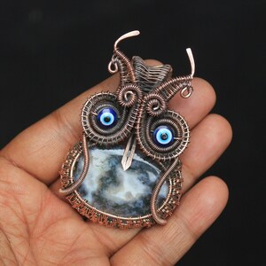 Może przedstawiać: Wisiorek owinięty miedzianym drutem z niebiesko-białym amuletem złego oka i szarym agatem.