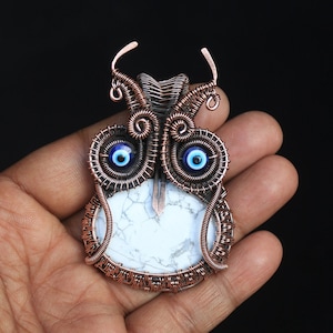 Może przedstawiać: Wisiorek w kształcie sowy z drutu miedzianego z białym kamieniem howlitu i niebieskimi koralikami złego oka.