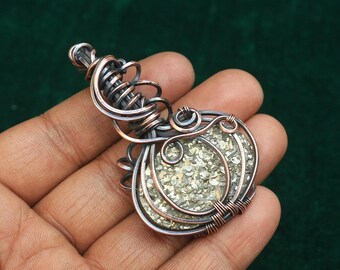 Könnte beinhalten: Ein Anhänger aus Silber- und Kupferdraht, der einen runden, hellgrauen Stein mit einer funkelnden, glitzernden Textur umwickelt.