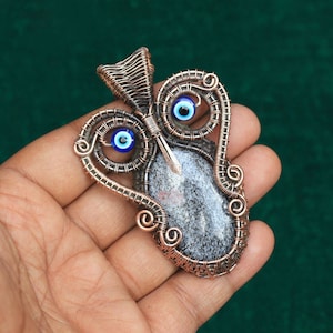 Könnte beinhalten: Ein mit Kupferdraht umwickelter Anhänger mit einem grauen und weißen Stein und zwei blauen Glücksbringer-Anhängern.