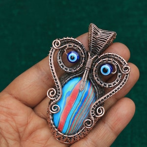 Może przedstawiać: Zawieszka owinięta miedzianym drutem z niebiesko-czerwonym marmurowym kamieniem i dwoma niebieskimi amuletami złego oka.