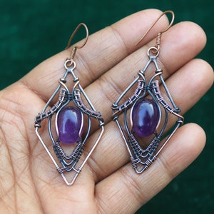 May include: A pair of amethyst gemstone earrings with a copper wire cage design. The earrings are a geometric shape with a purple stone in the centre.