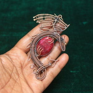 Puede incluir: Un colgante de dragón de alambre de cobre con una piedra preciosa roja. El dragón está enroscado alrededor de la piedra preciosa, con las alas extendidas.