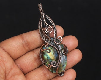 May include: A copper wire wrapped pendant featuring a green and blue labradorite gemstone. The pendant has a delicate, intricate design with swirls and loops.
