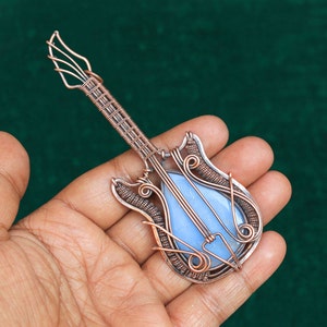 May include: A copper wire guitar pendant featuring a blue agate stone at its centre. The pendant is intricately crafted with wirework, making it a distinctive piece of jewellery.