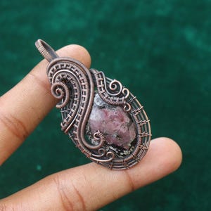 Könnte beinhalten: Ein tropfenförmiger Anhänger mit einem zentralen, rohen rosa-schwarzen Stein, der von einer komplexen Kupferdrahtwicklung umgeben ist. Die Metallarbeit besteht aus mehreren gewickelten und geflochtenen Kupfersträngen, die einen Rahmen bilden.