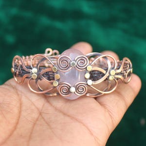 May include: A copper wire wrapped bracelet with a large, oval-shaped, light pink gemstone in the center. The bracelet is decorated with small, round, gold-colored beads.