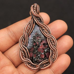 以下が含まれることがあります： 黒と赤の斑点のある石を特徴とする、銅線で包まれたペンダント。石は涙のしずくの形にセットされており、ワイヤーはねじれて装飾的なデザインに織り込まれています。
