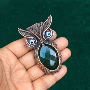 Może przedstawiać: Miedziany wisiorek w kształcie sowy z fasetowanym, ciemnozielonym korpusem z kamienia szlachetnego i niebieskimi koralikami w oczach. Skrzydła i głowa sowy są misternie tkane z drutu miedzianego, tworząc unikatową, ręcznie robioną biżuterię.