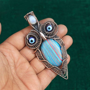 Może przedstawiać: Wisiorek w kształcie sowy owinięty miedzianym drutem z niebiesko-czerwonym paskowanym kamieniem i dwoma niebieskimi amuletami w kształcie oka.