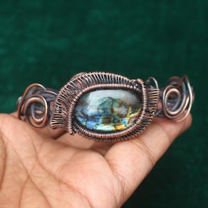 May include: A handcrafted copper wire bracelet featuring a central oval labradorite stone. The stone displays iridescent blue, green, and yellow hues. The bracelet's design incorporates intricate wire wrapping and decorative swirls.