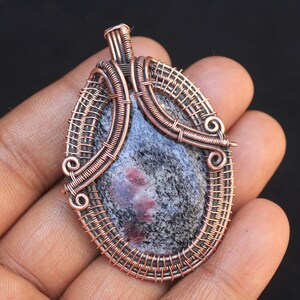 May include: A copper wire wrapped pendant featuring a gray and pink stone with red inclusions. The pendant is oval shaped and has a wire loop at the top for hanging.