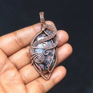 Peut inclure: Un pendentif en fil de cuivre enveloppé avec une pierre mouchetée noire et rose. Le pendentif a un design unique et complexe avec un motif en spirale.