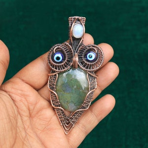 Może przedstawiać: Wisiorek w kształcie sowy owinięty miedzianym drutem z zielonym agatem i niebieskimi akcentami złego oka. Wisiorek ma księżycowy kamień na górze.