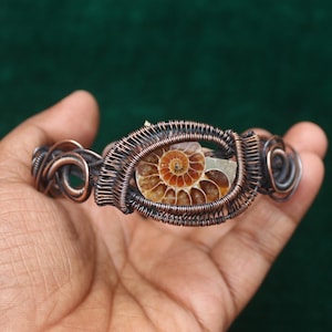 May include: A handcrafted copper wire bracelet featuring a fossilized ammonite center. The ammonite displays a spiral pattern in shades of brown and orange. The bracelet's wirework has a woven design.