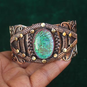 May include: A handcrafted copper cuff bracelet with an oval, iridescent green stone at the centre. The bracelet features intricate wirework and small gold-coloured accents, creating a unique, artistic design. The cuff is wide and has a vintage aesthetic.