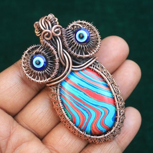 Może przedstawiać: Wisiorek owinięty miedzianym drutem z niebieskim i białym oczkiem i czerwono-niebiesko-zielonym marmurowym kamieniem.