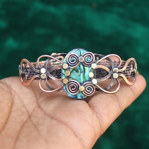 Puede incluir: Un brazalete de alambre de cobre con una gran incrustación de concha de abulón. El brazalete está decorado con pequeñas cuentas doradas y espirales de alambre.