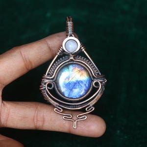 May include: A handcrafted pendant featuring a large, round, iridescent gemstone with blue, yellow, and orange hues, set within a copper wire frame. A smaller, round, white stone sits above the larger stone. The pendant has an intricate, openwork design.