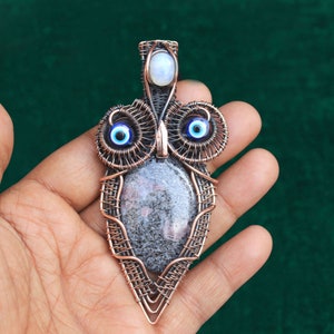 Może przedstawiać: Wisiorek w kształcie sowy owinięty miedzianym drutem z szarym kamieniem i niebieskimi akcentami złego oka.