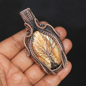 May include: A copper wire wrapped pendant featuring a tree agate stone with a brown, white, and orange pattern. The pendant is a unique piece of jewelry with a natural stone and wirework design.