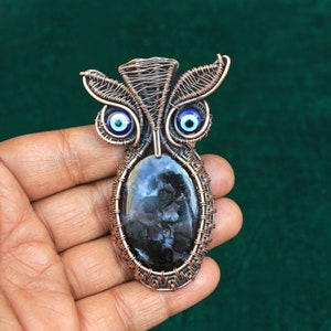 Może przedstawiać: Wisiorek w kształcie sowy z drutu miedzianego z czarno-białym agatem i niebieskimi koralikami złego oka.