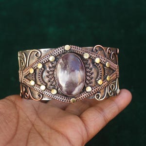 May include: A silver cuff bracelet with an oval, purple-hued stone at its center. The bracelet features intricate copper wirework and small gold-colored accents. The design is ornate and detailed, showcasing a blend of metals.