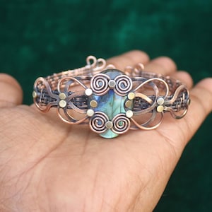 Peut inclure: Un bracelet en fil de cuivre enveloppé avec une grande pierre précieuse irisée de forme ovale au centre. Le bracelet présente un design en fil tissé avec des volutes décoratives et de petites accents dorés.
