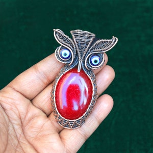 Może przedstawiać: Ręcznie robiony wisiorek w kształcie sowy z czerwonym owalnym kamieniem pośrodku. Wisiorek posiada miedziane detale z drutu, niebieskie akcenty w kształcie oka i wzór z drutu. Oczy sowy to małe niebiesko-białe szklane koraliki.