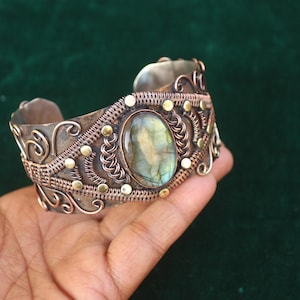 Puede incluir: Un brazalete de puño artesanal con una piedra central ovalada de labradorita. El metal plateado está adornado con intrincados trabajos de alambre, detalles en cobre y pequeños tachuelas doradas. El brazalete tiene una estética vintage o bohemia.