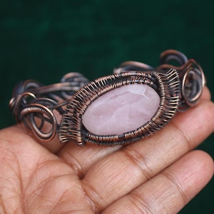 May include: A handcrafted copper wire bracelet featuring a central oval rose quartz stone. The intricate wirework surrounds the pink gemstone, creating a unique, artistic design. The bracelet is a piece of jewelry.