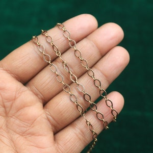 May include: Close-up of a delicate gold-colored chain with an oval link design. The chain is held against a dark green background, showcasing its intricate details and warm metallic hue. Ideal for jewelry making or crafting.