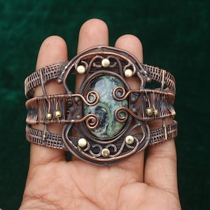 May include: Copper wire-wrapped bracelet with an oval, green stone center. The bracelet features intricate wirework with gold-colored accents. The design includes swirls and geometric patterns, creating a unique, handcrafted look.