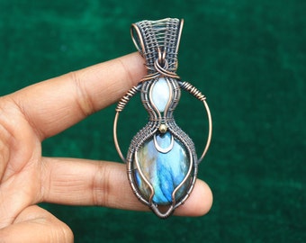 May include: A wire-wrapped pendant featuring a blue labradorite gemstone, a white moonstone, and copper wire. The pendant is held by a finger against a green background.