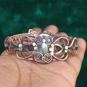 May include: A copper wire bracelet with a gray stone and gold accents. The bracelet is woven with a delicate pattern and features a large, oval-shaped stone in the center.