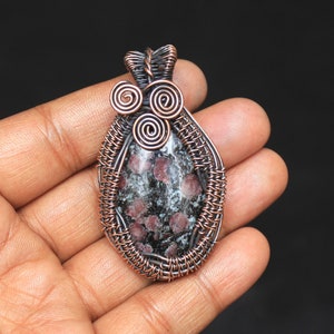 Pode incluir: Um pingente envolto em fio de cobre com uma pedra preciosa preta e branca com inclusões vermelhas. O pingente tem um design único com três espirais de fio de cobre na parte superior.