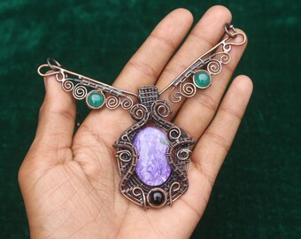 May include: A handcrafted copper wire necklace featuring a central pendant with a purple stone and a black stone accent. The necklace has green stone accents and decorative wirework. The design is held in a hand.