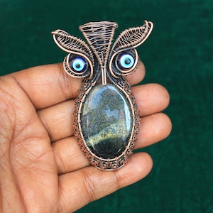 Może przedstawiać: Ręcznie wykonany wisiorek w kształcie sowy z drutu miedzianego z centralnym owalnym kamieniem. Sowa ma niebieskie szklane oczy i skomplikowane detale z drutu, tworząc unikalny, artystyczny element. Kamień ma mieszankę kolorów zielonego, czarnego i żółtego.