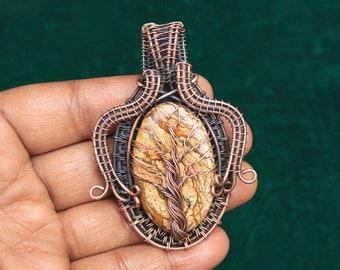 May include: A handcrafted pendant featuring an oval-shaped stone with a tree design, encased in copper wire. The pendant has a brown and tan stone with a detailed tree motif. The wirework is intricate, with a mix of copper and darker metal.