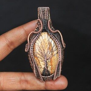 May include: A copper wire wrapped pendant featuring a tree agate stone with a natural tree pattern. The pendant is oval shaped and has a copper bail.
